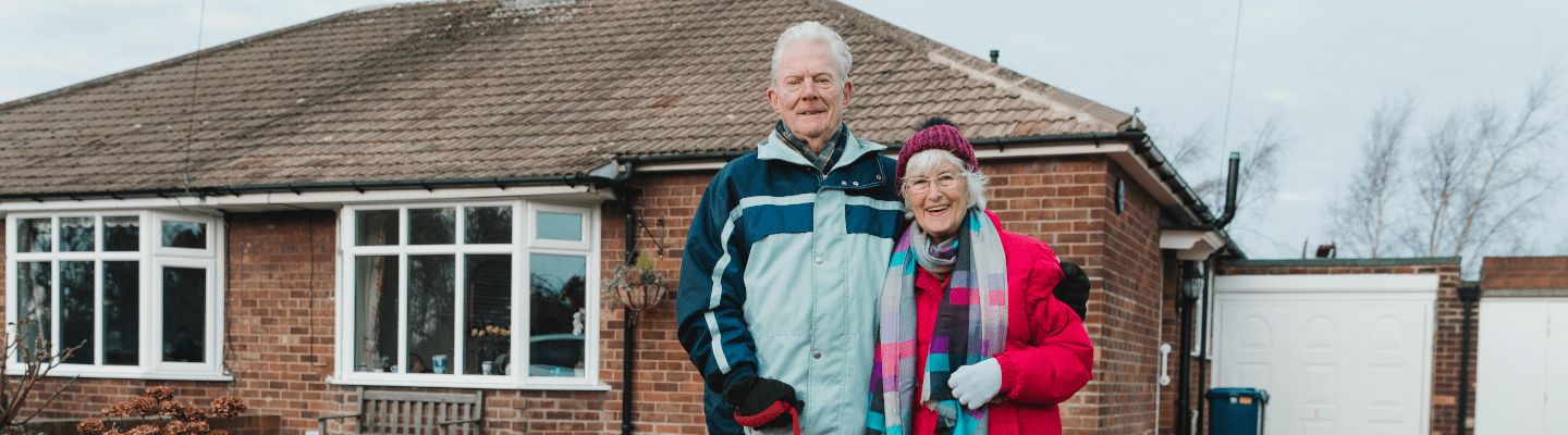 An elderly couple stands outside their bungalow